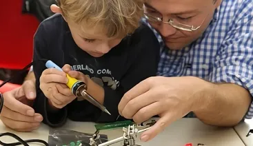 Pai ajudando o filho a usar um ferro de solda em cima da mesa do hackerspace
